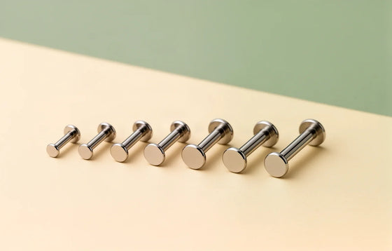 A row of titanium flat-back labret bars in different gauges and lengths on a cream surface