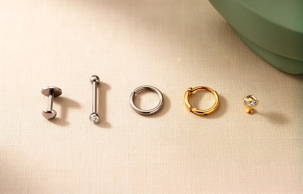 Piercing jewellery types laid out: labret, barbell, seamless ring, clicker and stud in titanium and gold