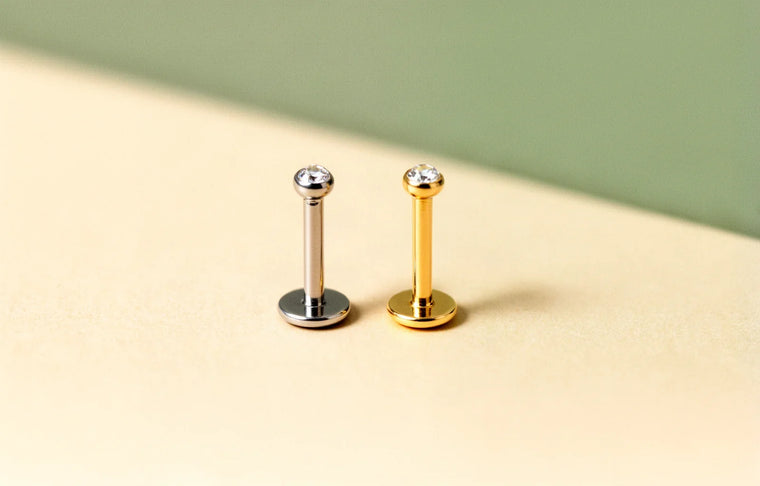 A titanium and a gold flat-back piercing stud side by side on a cream surface
